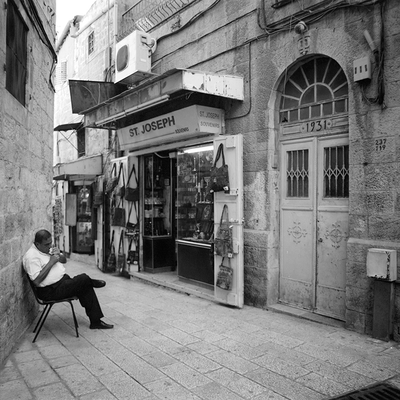 Black and white square photo of man sitting in an alleyway in the old city of Jerusalem