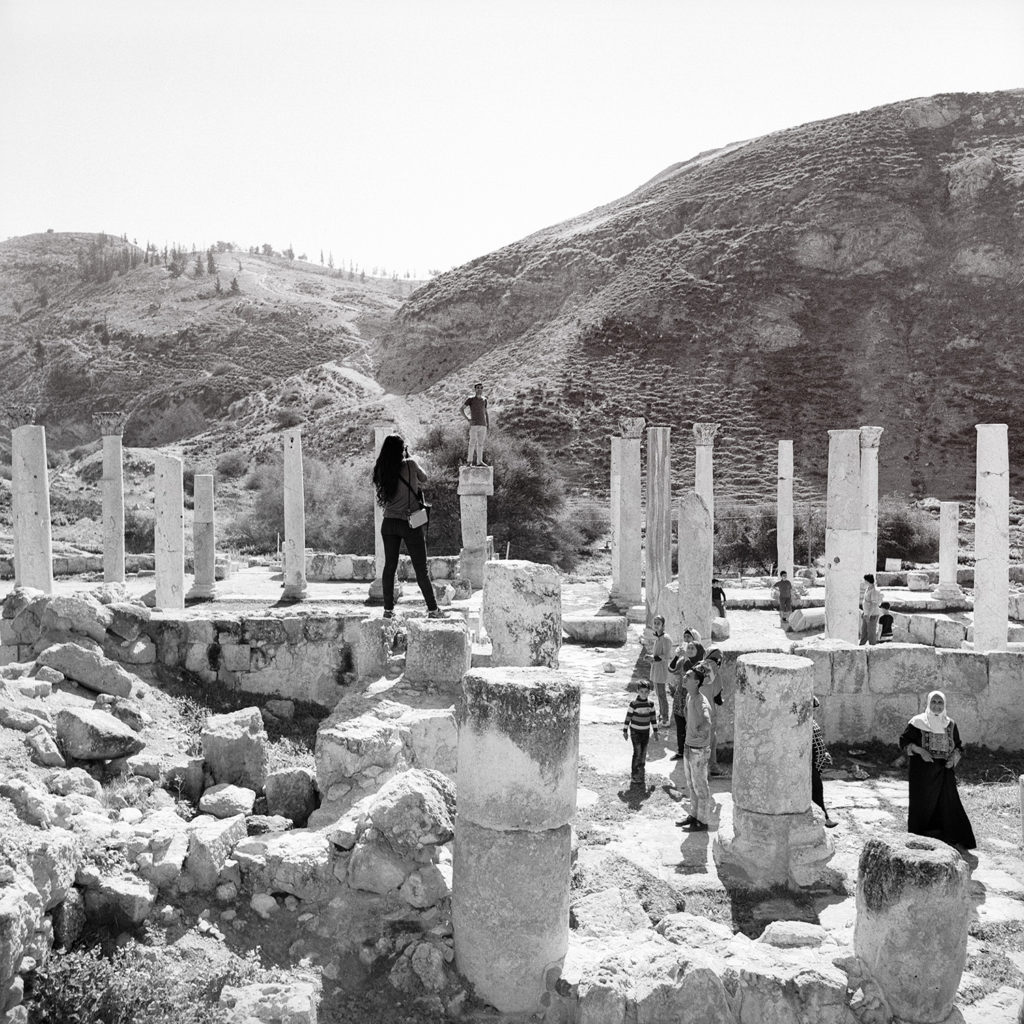 Black and white square photo of photographer standing on old Roman ruins taking a portrait with onlookers watching