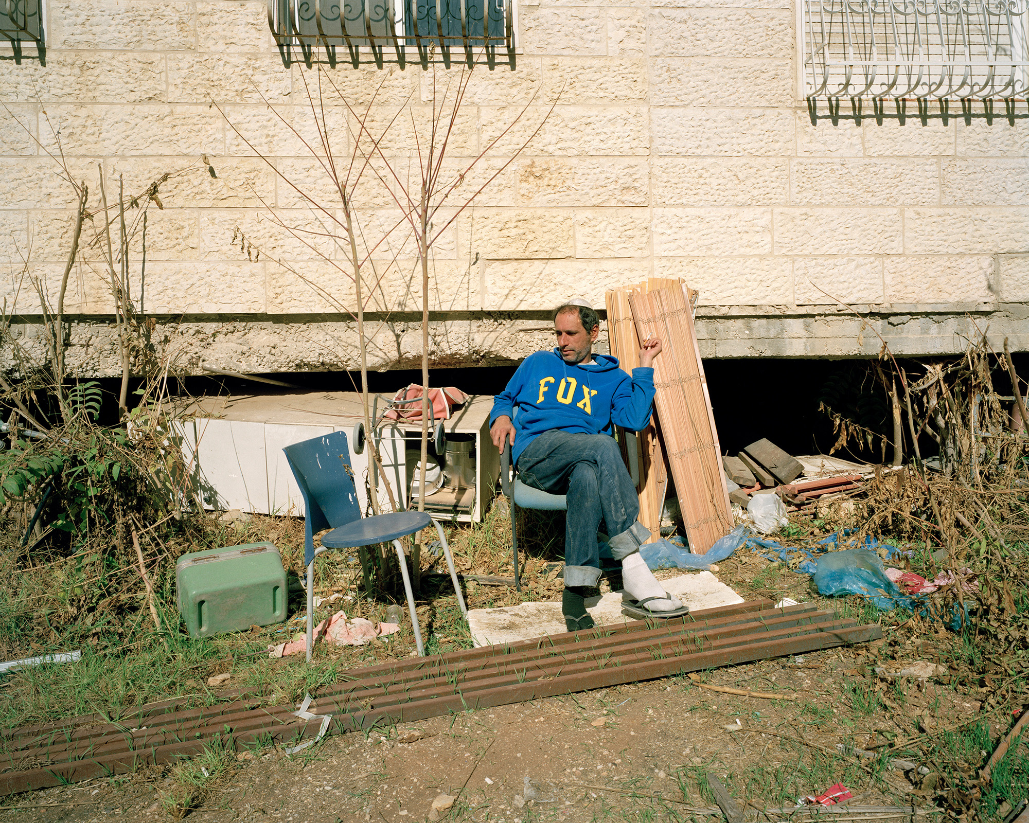 Man sitting in sun behind apartment building surrounded by debris